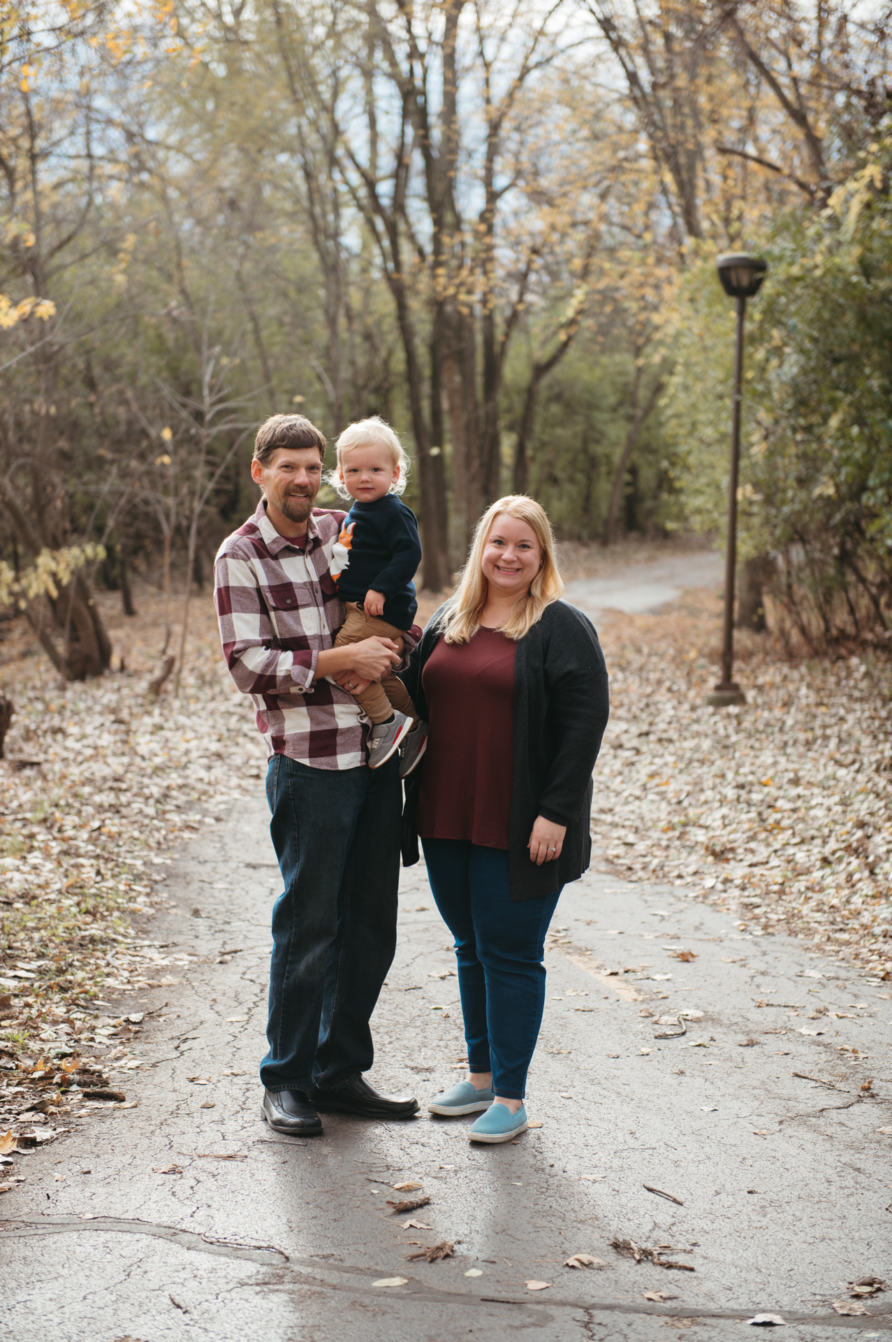 Dad holds toddler in fall pathway while mom smiles along nearby