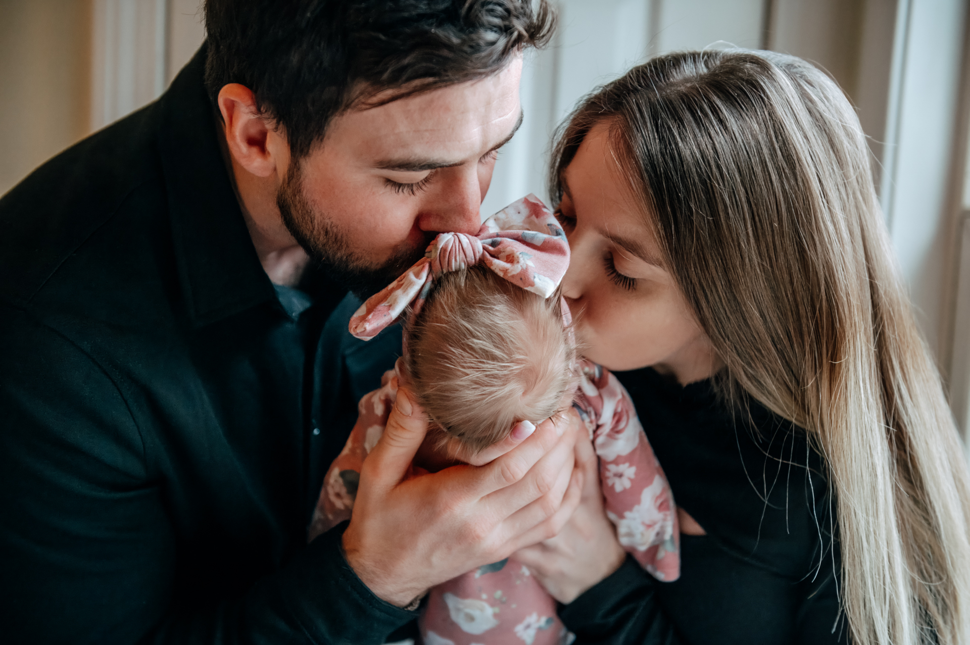 A mom and dad kiss the top of a baby girls head.