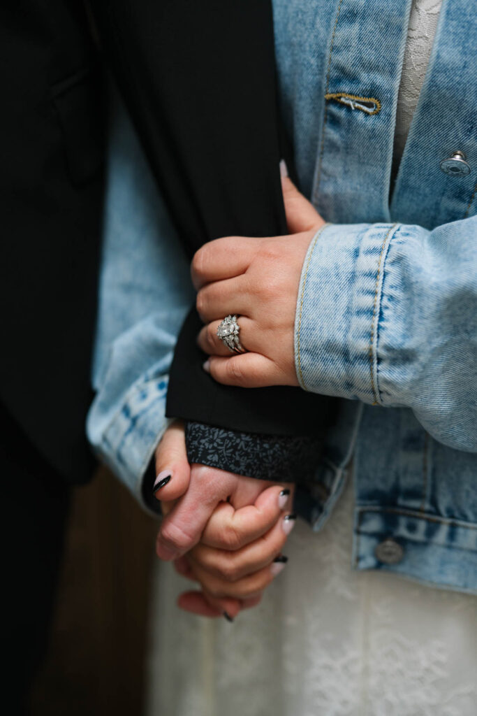 Close-up of two brides holding hands, showing engagement and wedding rings, one wearing a pearl-studded denim jacket