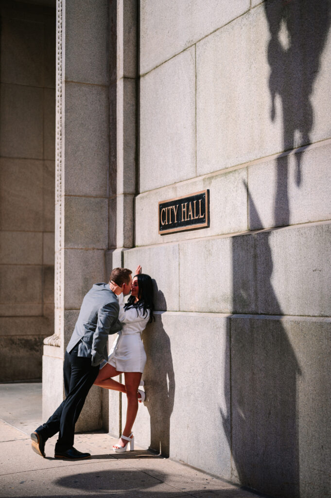 Bride in white tuxedo dress and groom in grey suit kiss against the limestone wall beneath the City Hall sign at their Chicago City Hall elopement