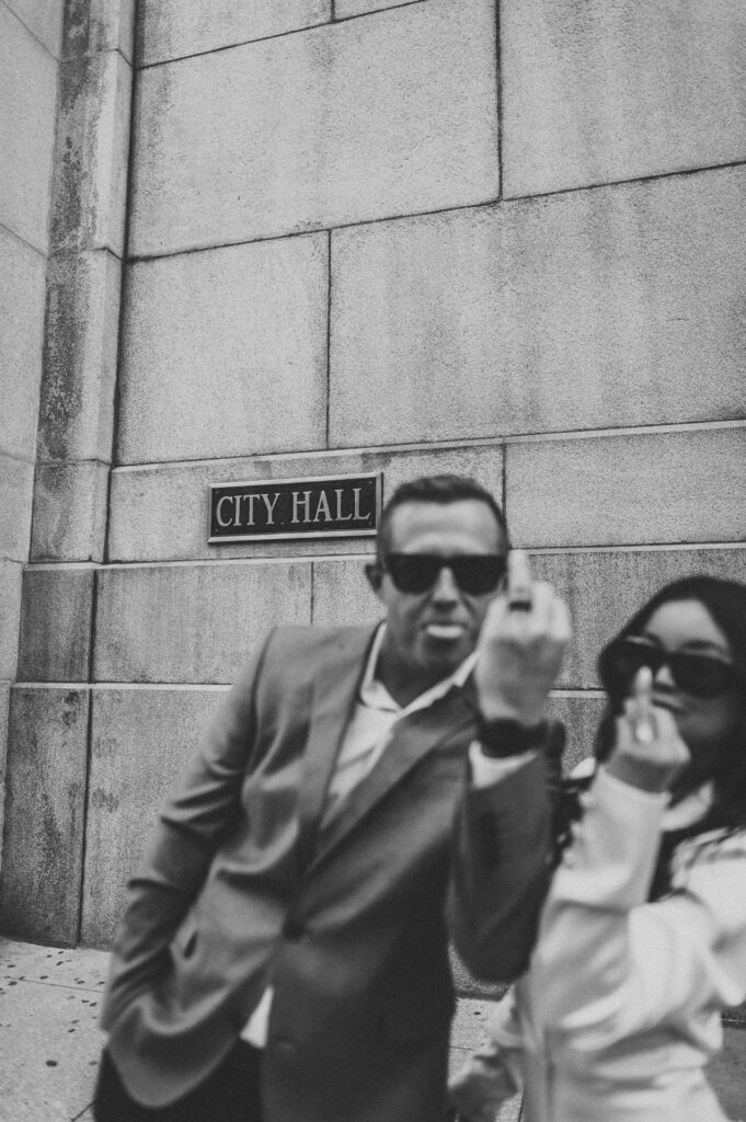 Black and white candid of bride and groom in sunglasses outside Chicago City Hall, the couple giving a playful ring finger to the camera