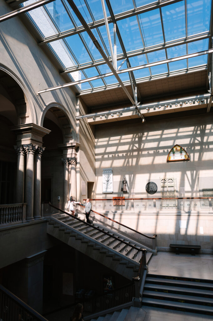 Wide architectural shot of bride and groom descending the grand staircase inside the Art Institute of Chicago, lit by overhead skylights